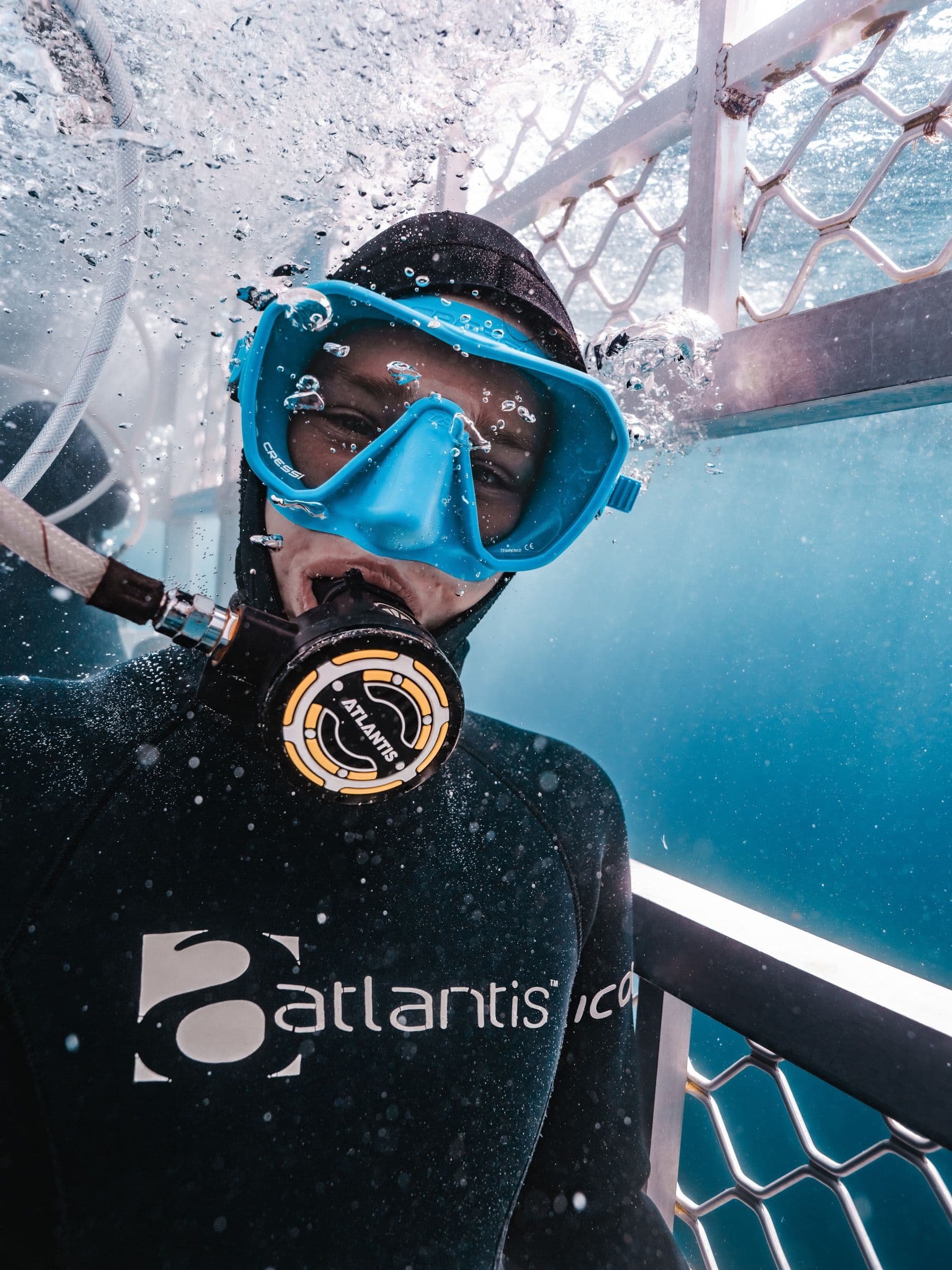 Diver in a wetsuit and blue mask breathes through a regulator inside a shark cage, with bubbles rising overhead.