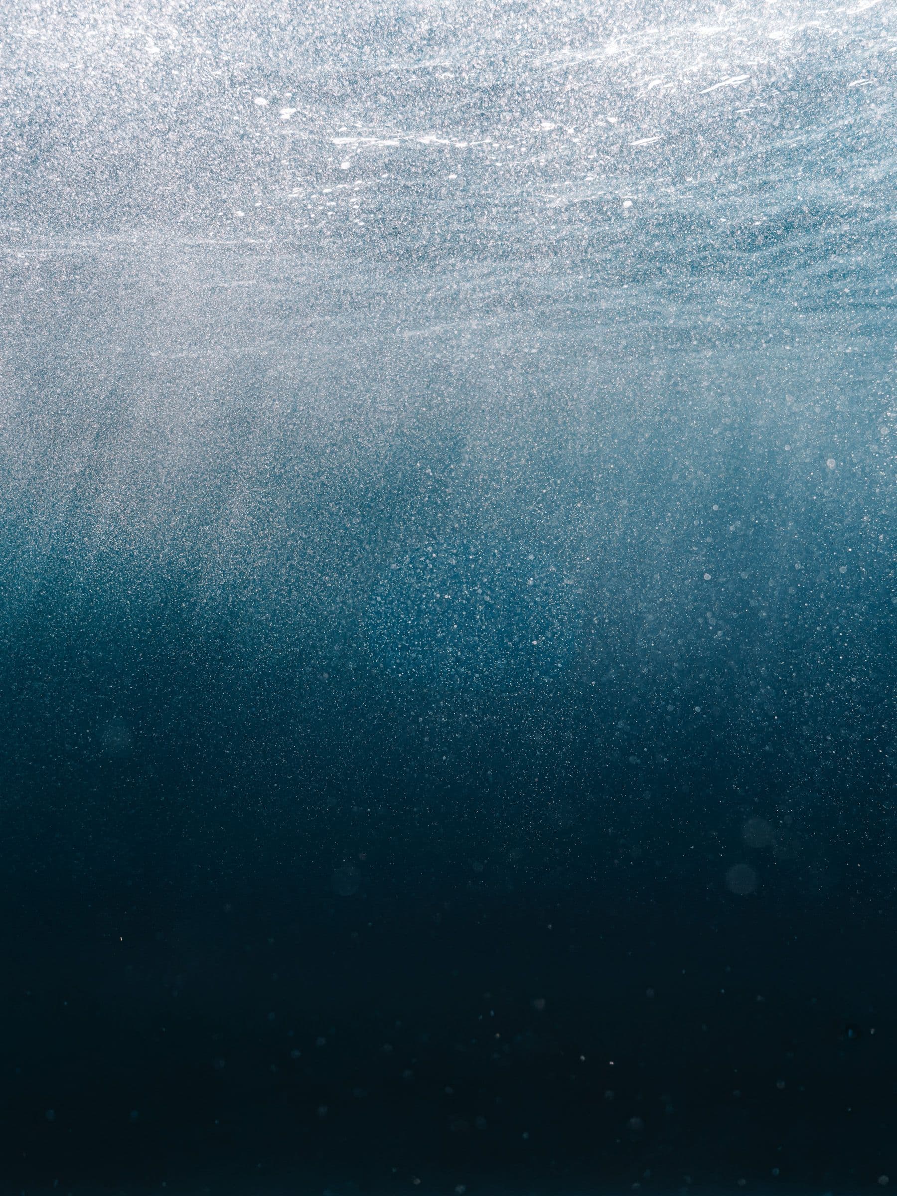 Underwater view looking up toward the surface, with sunbeams filtering through deep blue water and tiny particles.