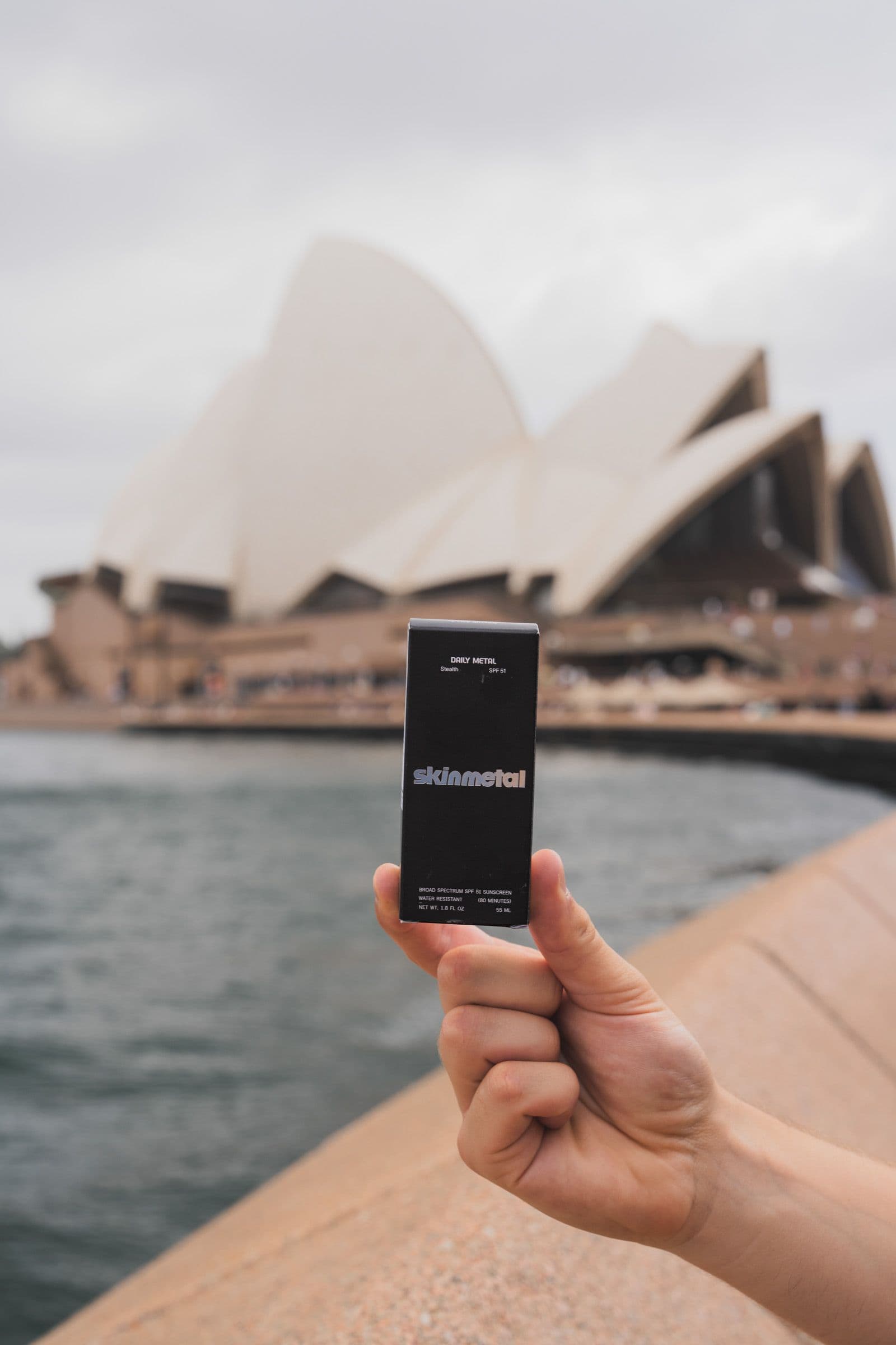Hand holds black Skinmetal sunscreen box with Sydney Opera House blurred in background.
