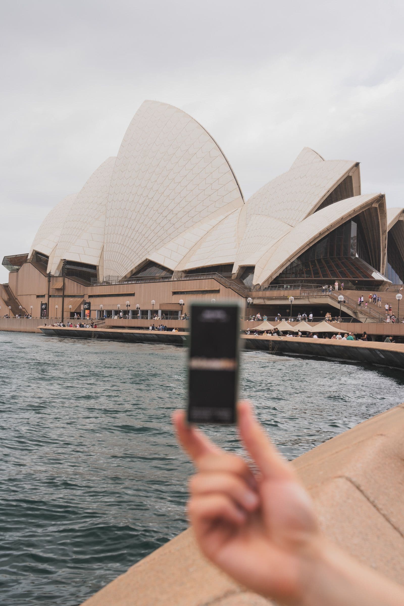 Hand raises a small black product box with the Sydney Opera House behind gray harbor water.