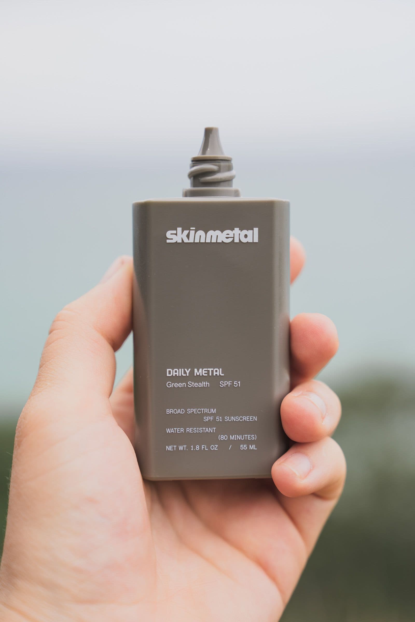 Hand presents a gray Skinmetal Daily Metal SPF 51 sunscreen bottle against ocean and sky.