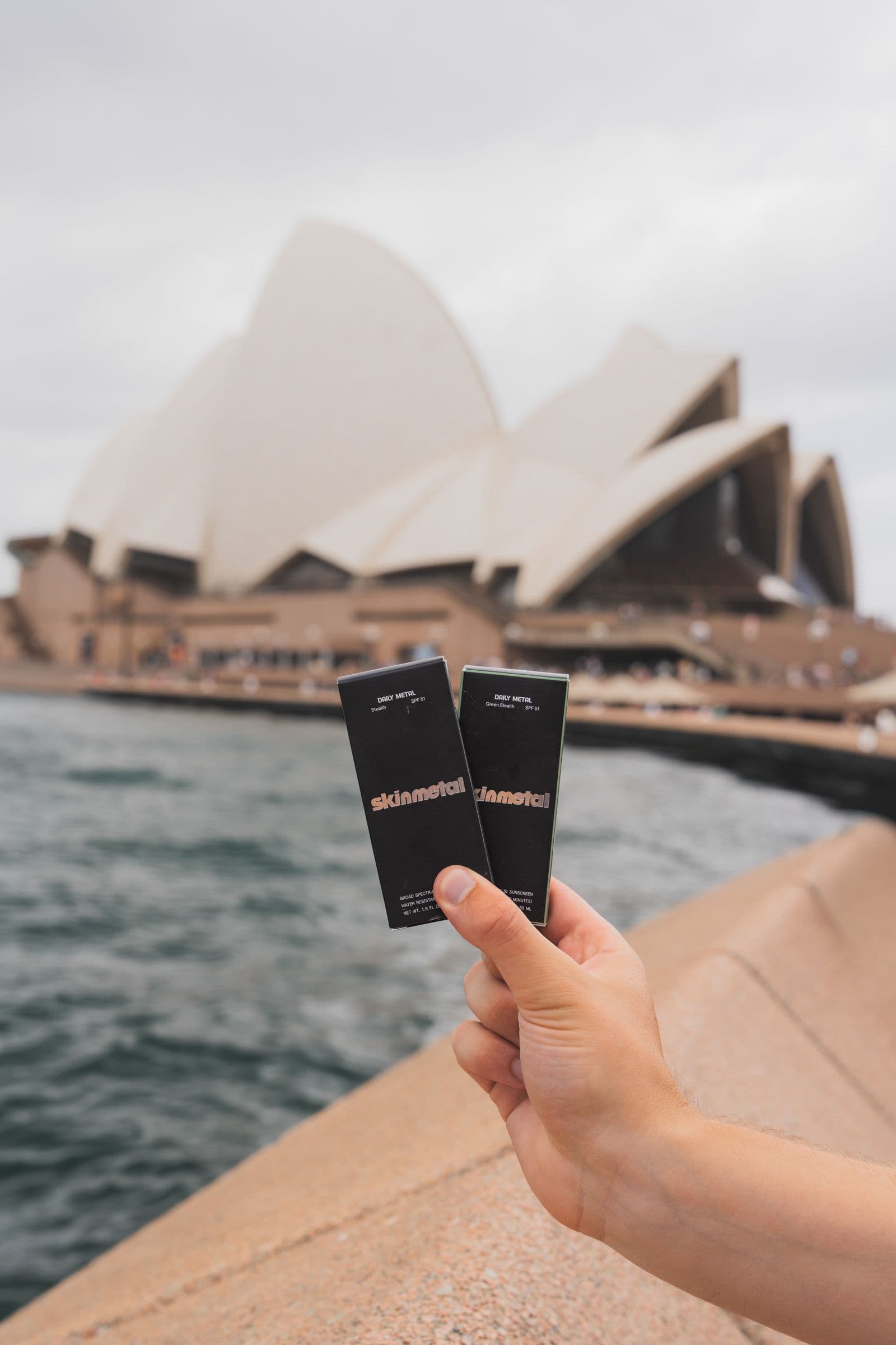 Hand holds two black Skinmetal sunscreen boxes side by side with Sydney Opera House behind.