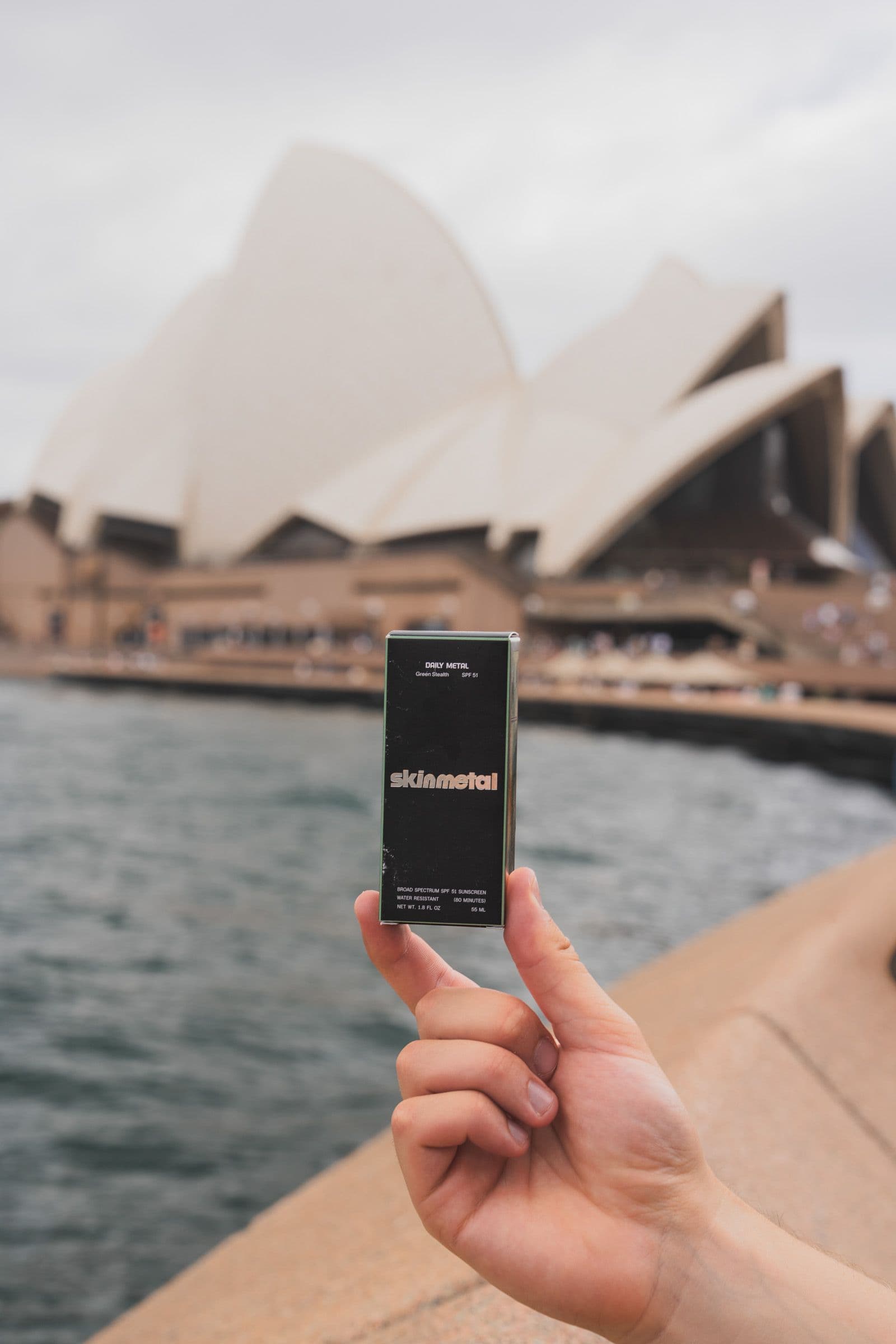 Hand holds black Skinmetal Green Stealth sunscreen box with Sydney Opera House in soft background.