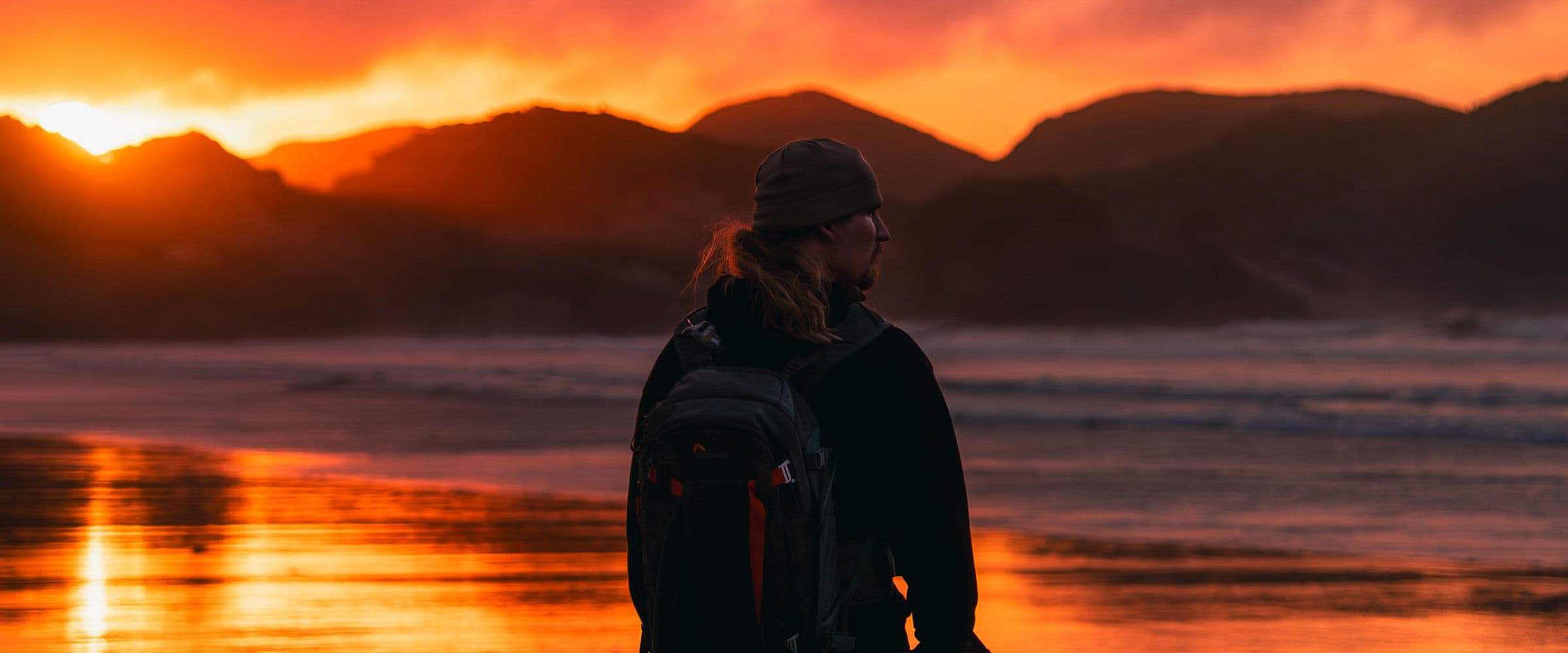 Silhouetted hiker with backpack and beanie watches a fiery orange sunset over coastal hills.