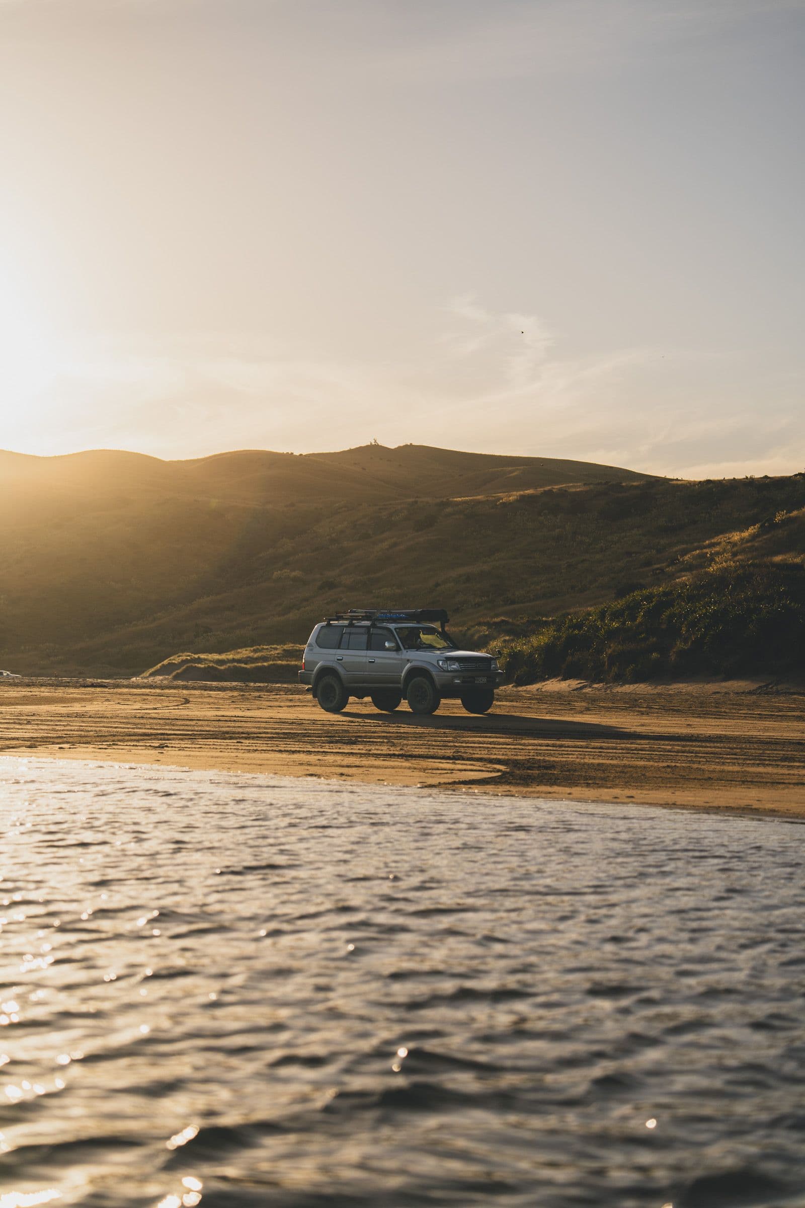 Silver four-wheel drive parked on a wide beach beside a tidal stream with hills beyond.