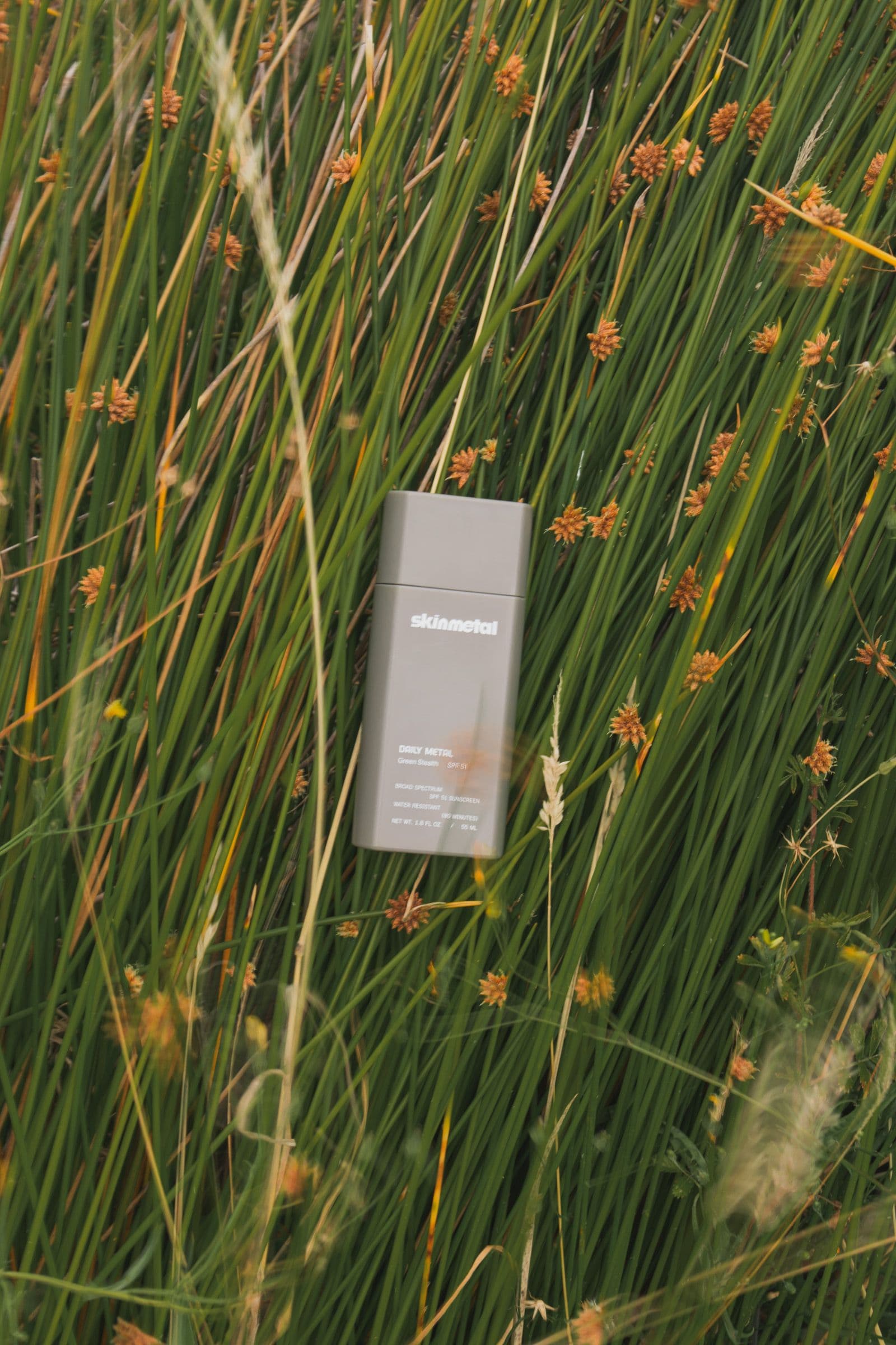 A gray Skinmetal sunscreen rests among tall green grass and small orange blooms.