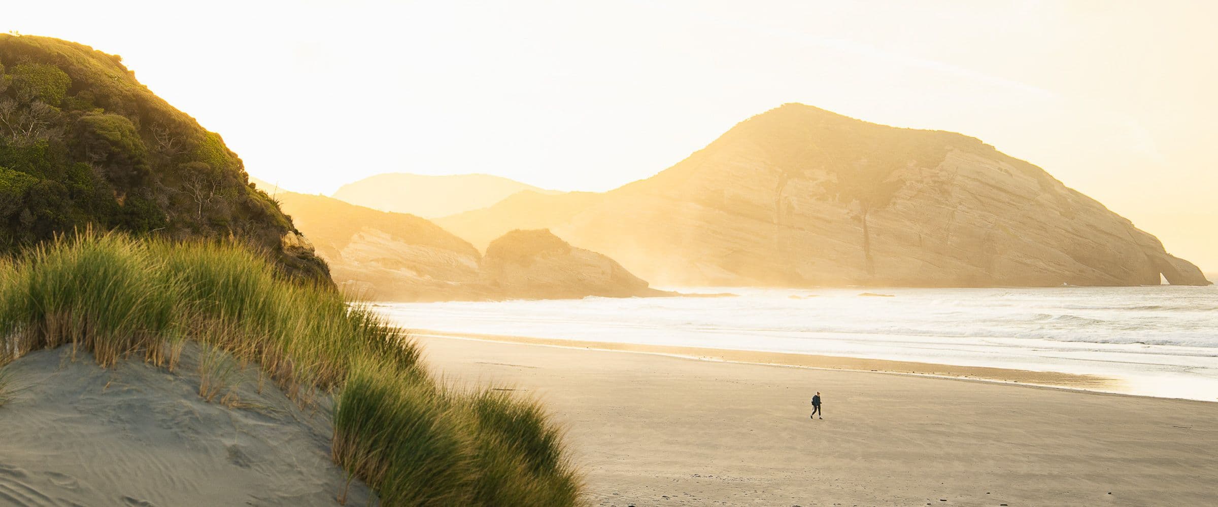 Tiny figure walks along wide sandy beach at sunset with grassy dunes and mountains glowing in haze.