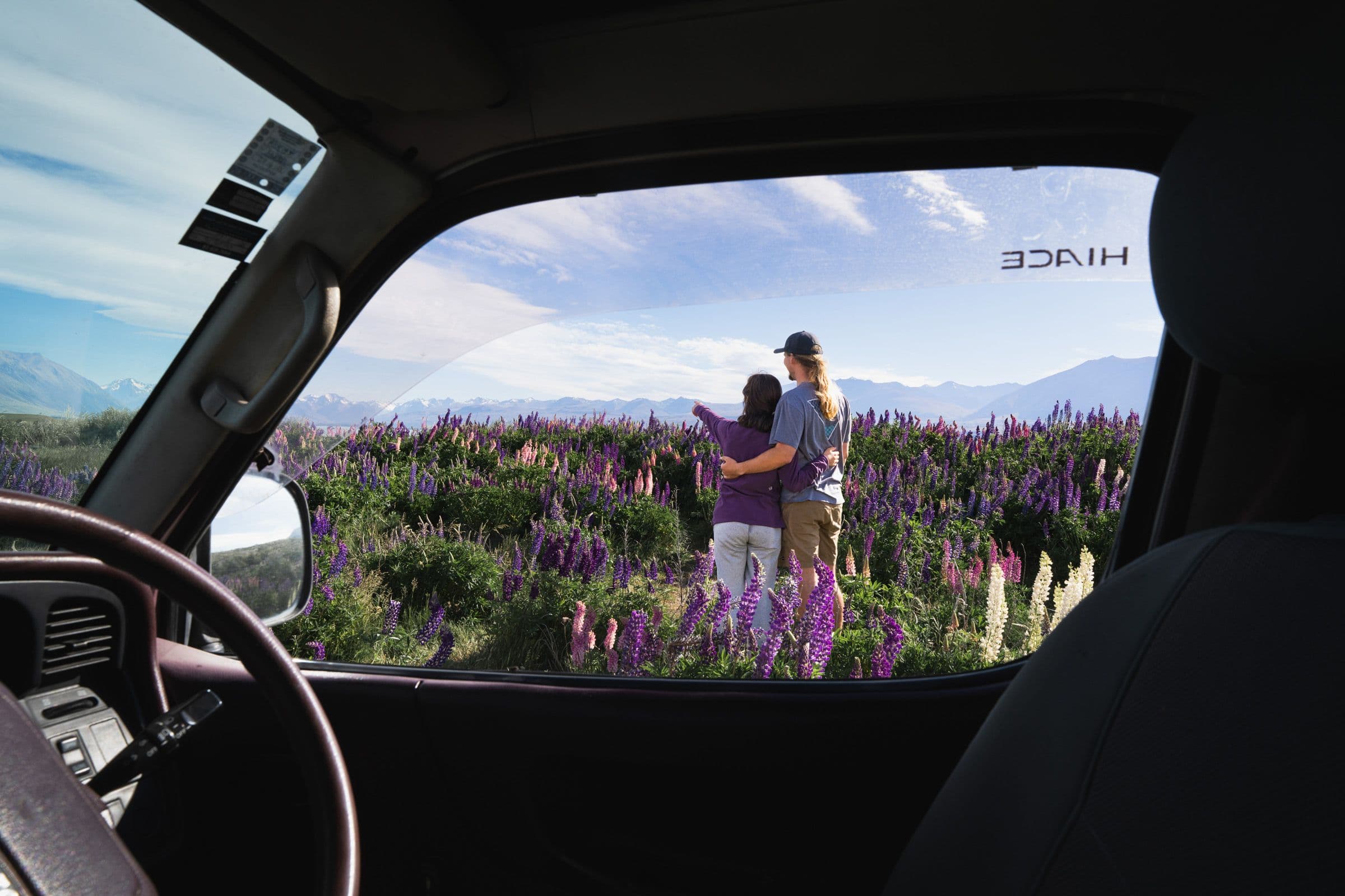View from inside a van of a couple embracing in a field of purple lupine flowers with mountains behind.