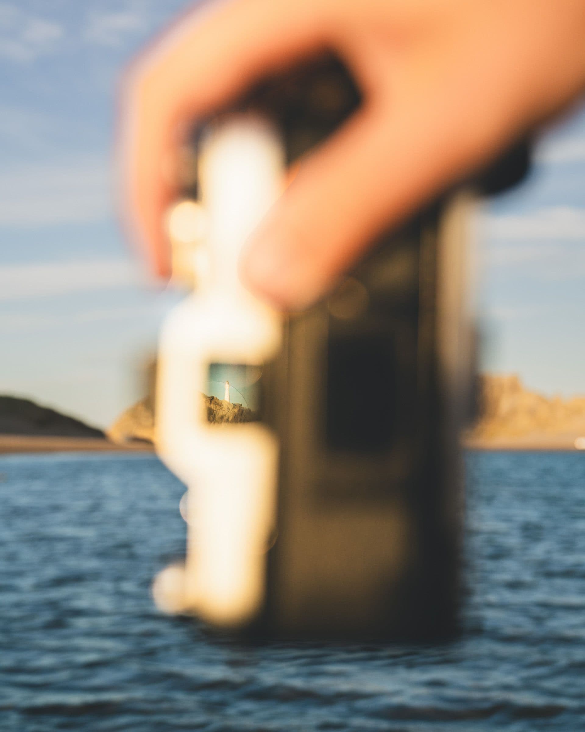 A hand holds a small glass bottle in front of a distant white lighthouse seen through the glass.