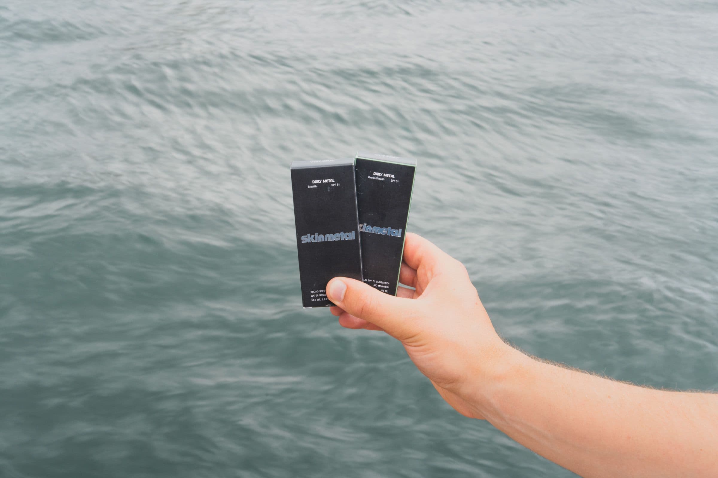 Hand holds two black Skinmetal sunscreen boxes above rippled gray-green ocean water.