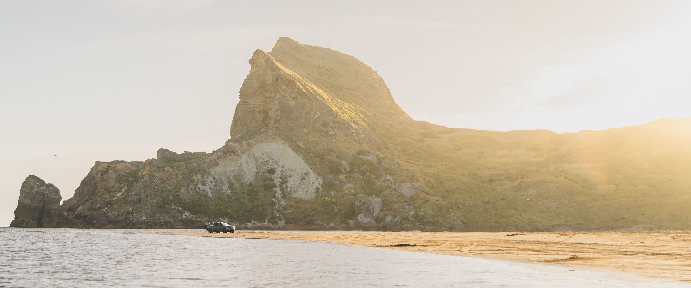 A small truck sits on a sandy spit beneath a massive green and gold headland catching sunlight.