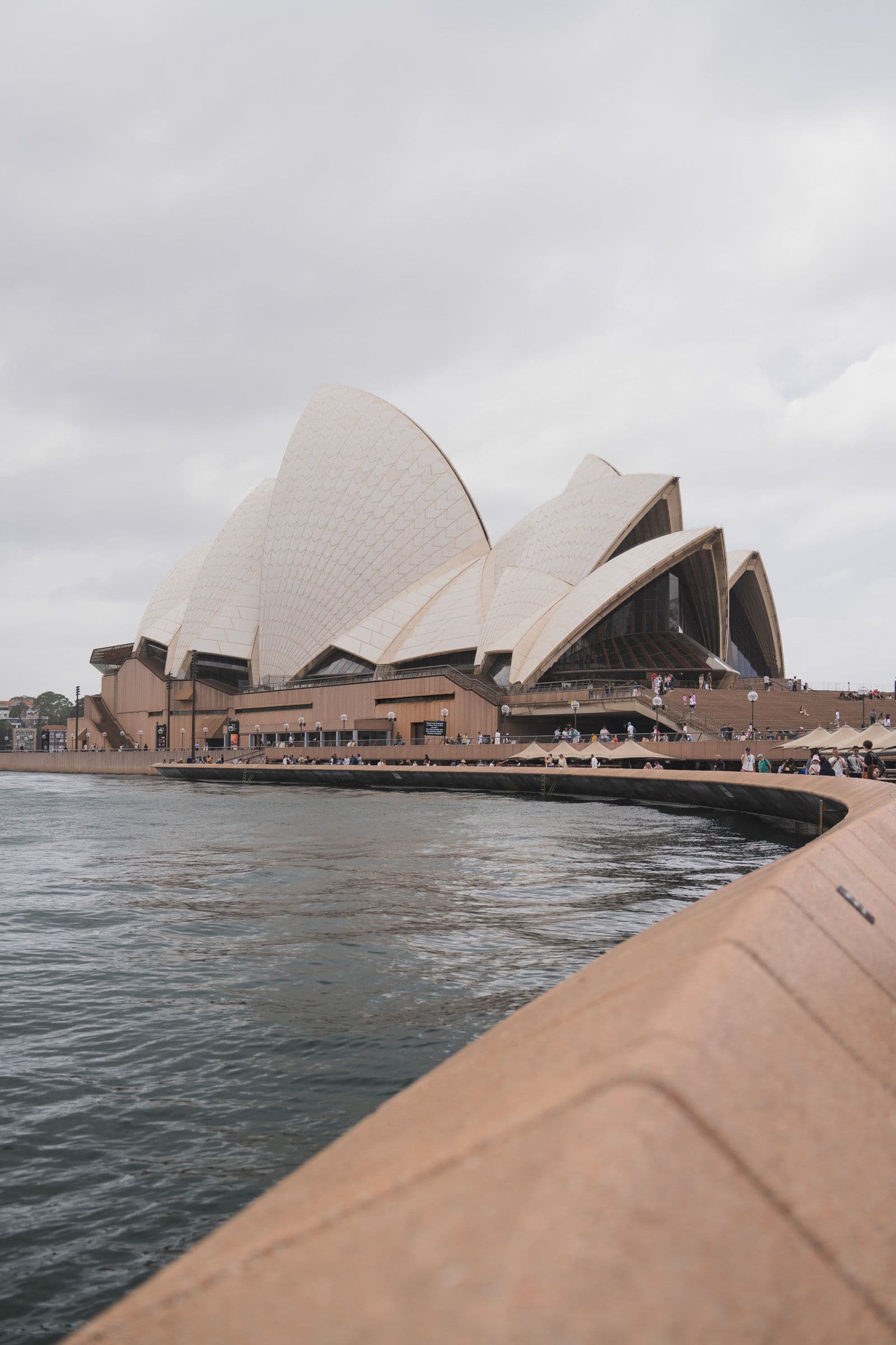 The Sydney Opera House and its white sail roofs seen from the waterfront under an overcast sky.