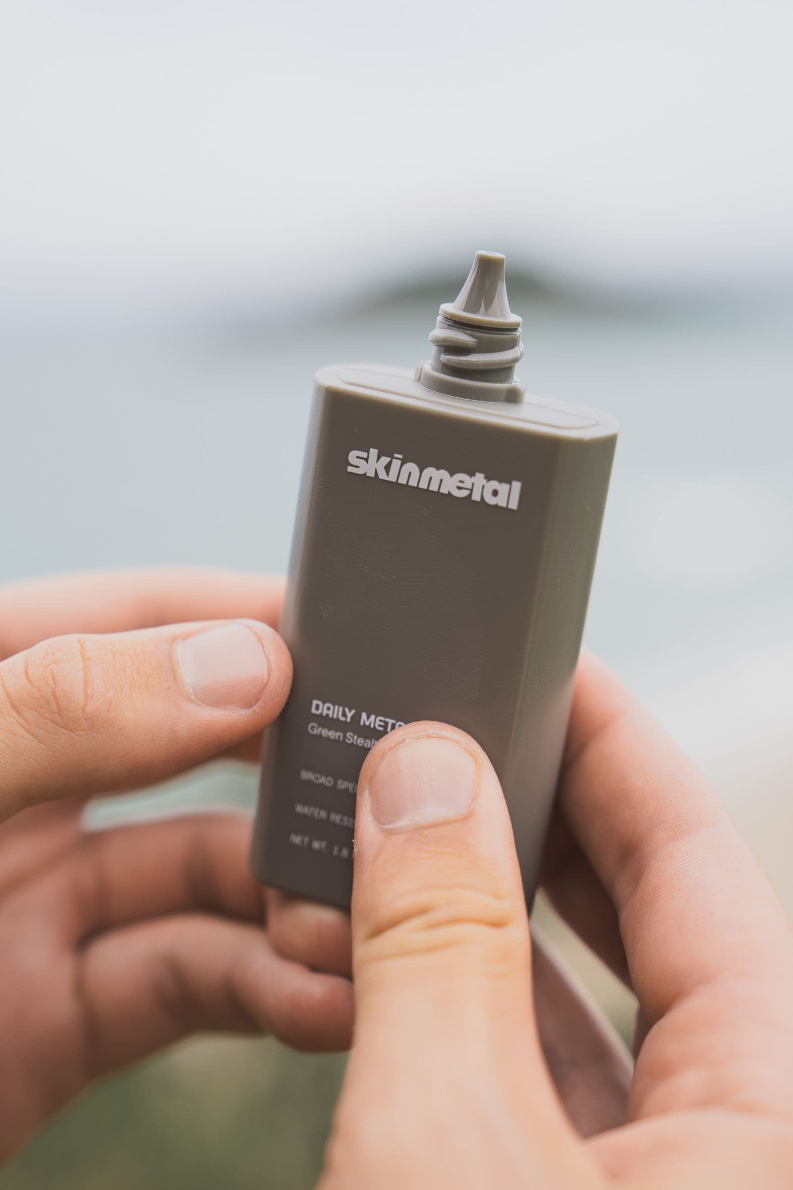 Hands hold a gray Skinmetal sunscreen bottle against a soft coastal background.