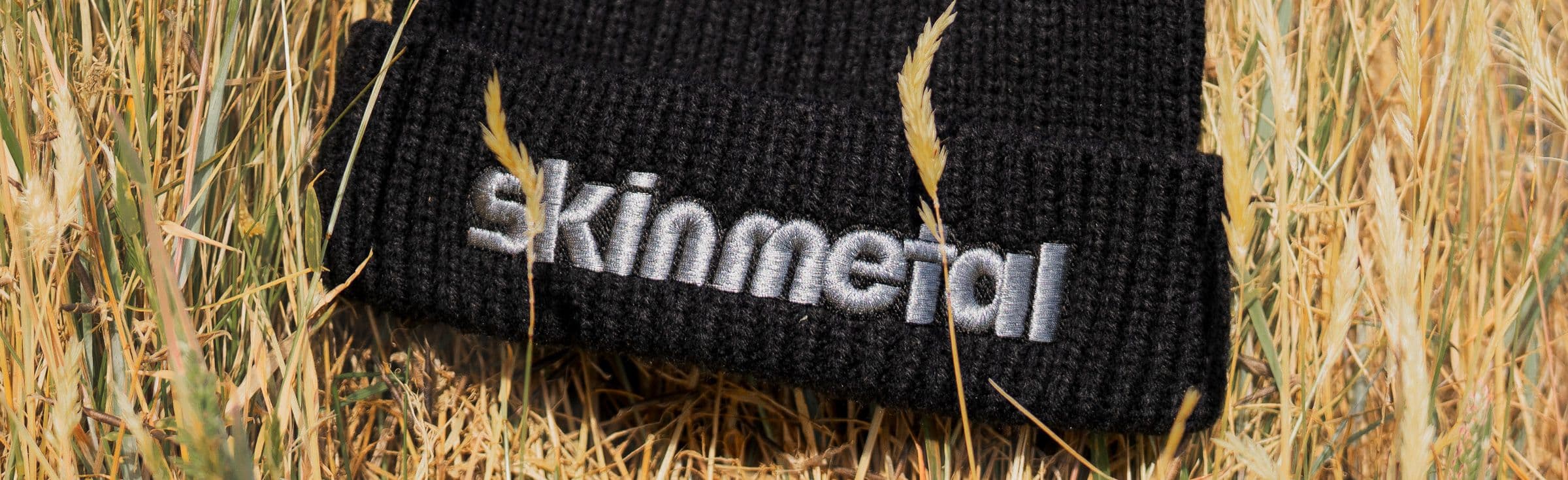 Black knit beanie embroidered with the word Skinmetal lying on dry golden grass.