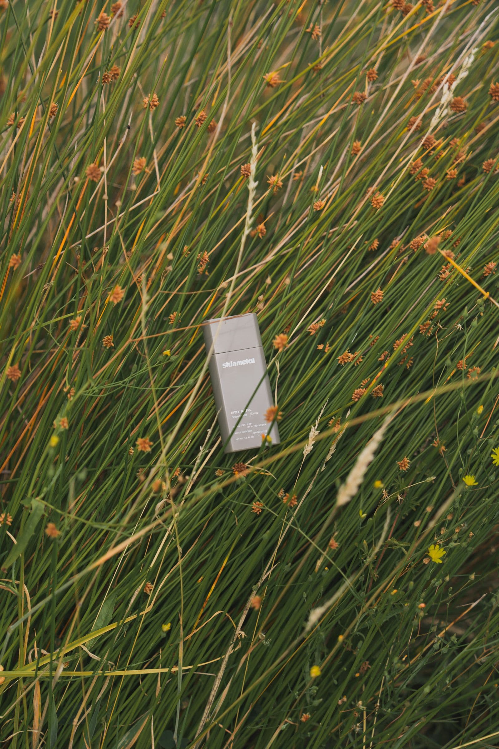 A gray Skinmetal sunscreen bottle nestled among tall green grass and wild flowers.