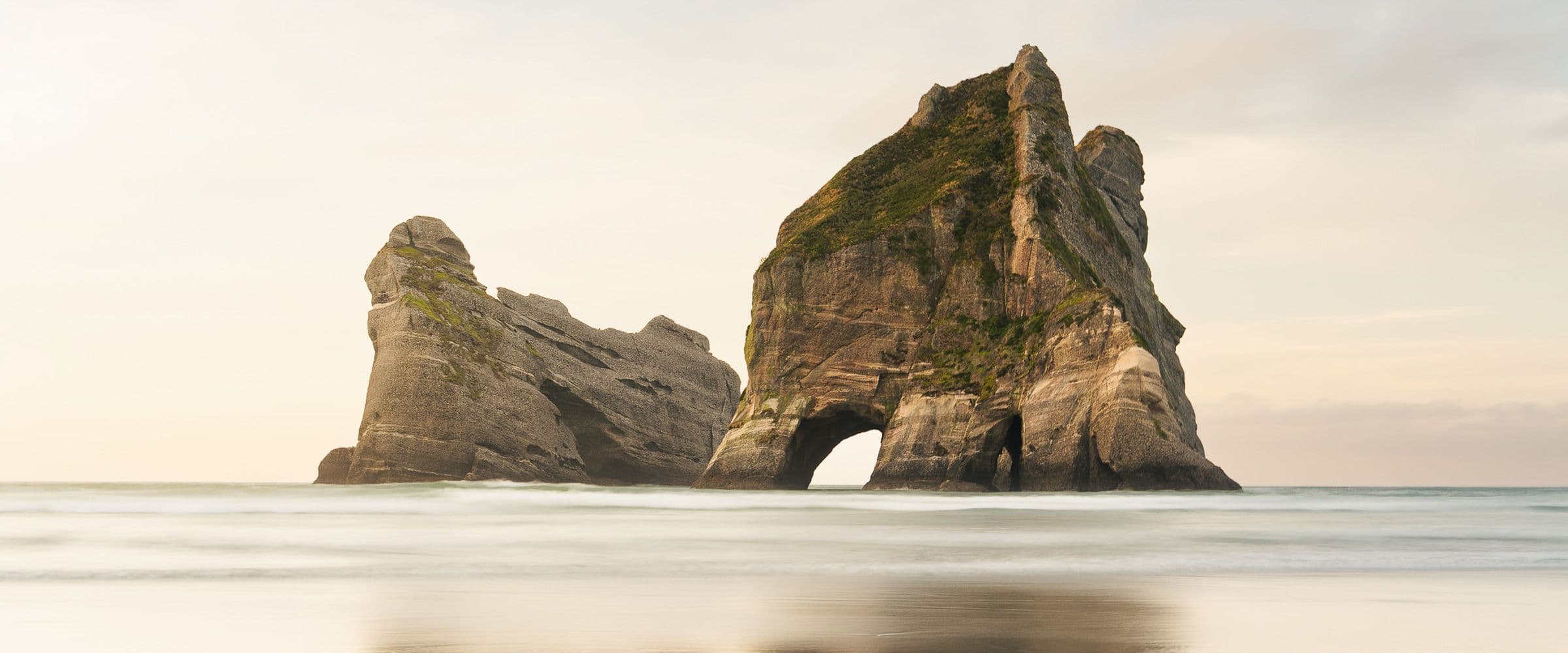 Twin sea stacks with a natural arch rise from calm water under a pale, overcast sky.
