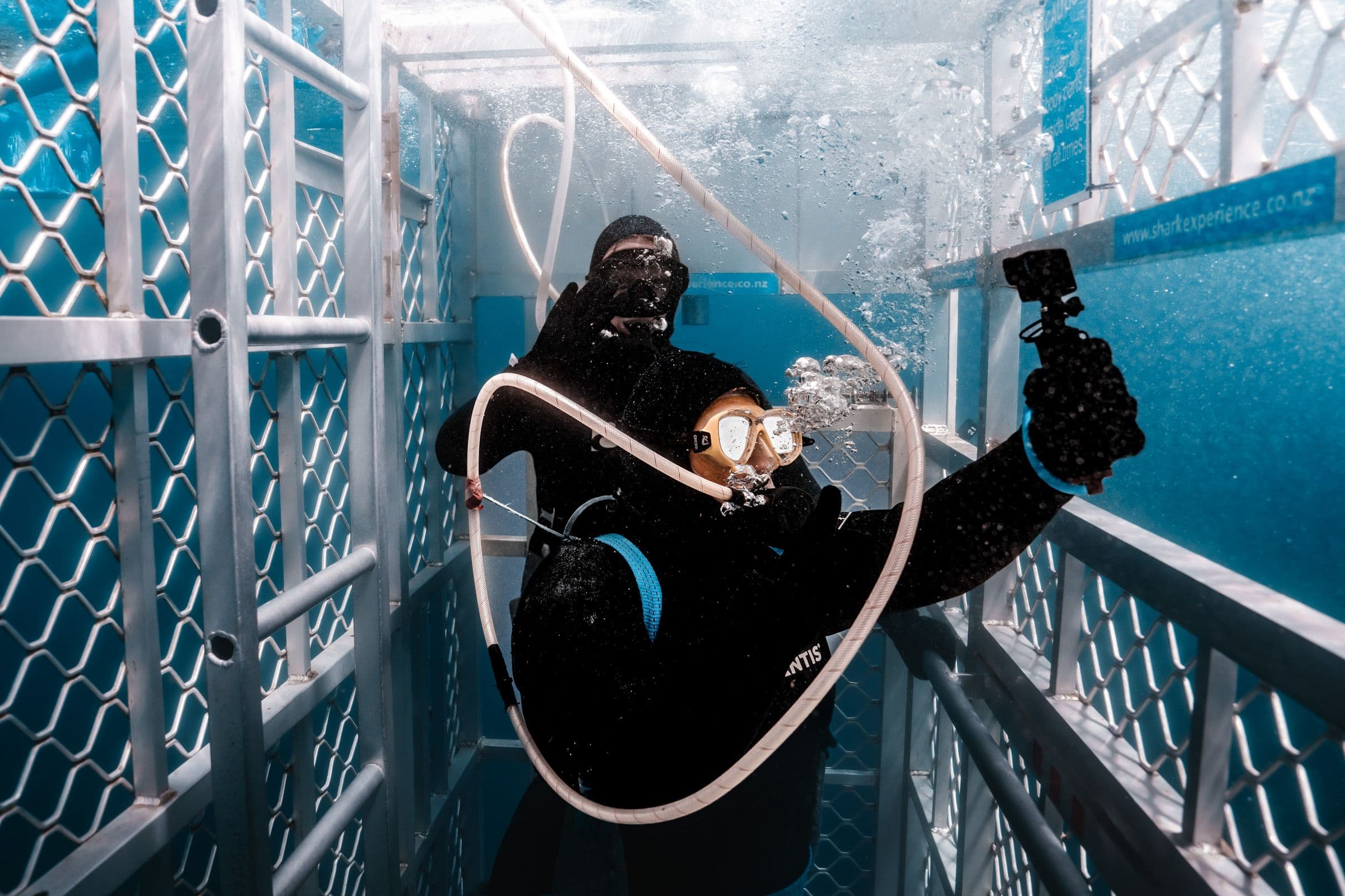 Diver in wetsuit holds a camera and regulator hose inside a metal shark cage, bubbles swirling around them.